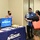 A NAVSUP (Naval Supply Systems Command) Fleet Logistics Center Pearl Harbor recruitment or informational booth is set up at what appears to be a job fair or community event. Four individuals are present, with a representative in a blue Hawaiian-style shirt reviewing documents with a visitor carrying a red tote bag. All participants are wearing face masks, indicating this event took place during the COVID-19 pandemic period.