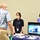 A professional event or job fair setting where two women are staffing an informational booth for 'Code 430 – Operations,' speaking with an older male visitor. The booth features a monitor displaying naval/military operational imagery and a blue-draped table with various materials. All three individuals are wearing face masks, suggesting this was taken during the COVID-19 pandemic period.