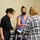 Three individuals are gathered around a table covered with a blue tablecloth, appearing to review documents in what looks like an indoor event or administrative setting. Two of the three people are wearing face masks, suggesting this was taken during or after the COVID-19 pandemic period. The interaction appears professional or administrative in nature, with papers being examined.