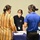 Three individuals are gathered around a table covered with a blue tablecloth at what appears to be an informational booth or conference event. A representative wearing a dark polo shirt with a lanyard and face mask is speaking with two visitors, one in a yellow striped blouse and one in a blue polo shirt. A monitor displaying a presentation slide partially visible with the word 'Contracting' is positioned on the table.