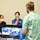 Three individuals are gathered around a table at what appears to be an informational booth or job fair. Two women behind the table, both wearing face masks, are presenting to a man in a Hawaiian shirt whose back is to the camera. A monitor on the table displays a slideshow with the text 'Community Involvement' and 'Care &amp; Support', associated with NAVSUP (Naval Supply Systems Command).