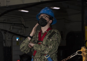 A U.S. Navy sailor wearing Navy Working Uniform (NWU Type III) is standing in what appears to be a ship's hangar bay or lower deck area. The sailor is equipped with a blue hard hat, black face mask, safety glasses, and a red and blue fall protection harness. The individual appears to be giving hand signals, possibly directing equipment or personnel movement.