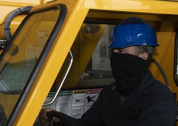 A worker is seated in the cab of a yellow industrial vehicle, likely a forklift or heavy equipment machine. The individual is wearing a blue hard hat, dark face covering, and navy blue coveralls or work uniform. Safety equipment including a fire extinguisher is visible inside the cab.