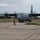 Ramstein Airmen enable Agile Spirit A U.S. Air Force C-130J Super Hercules military transport aircraft is being marshaled by a ground crew member on a concrete taxiway or apron. All four turboprop engines are running, with propellers visibly spinning. The setting appears to be a somewhat aged or austere airfield with cracked concrete and overgrown vegetation.