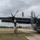 Ramstein Airmen enable Agile Spirit Military personnel are performing maintenance on a large gray military transport aircraft, identified by the visible US flag insignia as an American military plane, likely a C-130 Hercules variant. Three service members are working on the left wing area, with one standing on top of the wing, one climbing a ladder, and one at the base of the ladder. The scene takes place on a military airfield with overcast skies and a helicopter visible in the background.
