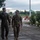 Ramstein Airmen enable Agile Spirit Two military personnel in the foreground are walking together on a wet paved surface, appearing to be in conversation. One wears a dark olive flight suit with a wing insignia patch, consistent with a US Air Force pilot, while the other wears a multicam uniform with what appears to be a Georgian flag patch. Several other uniformed personnel are visible in the background near what appears to be a military installation entrance or checkpoint.