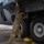 Ramstein Airmen enable Agile Spirit A military service member in OCP (Operational Camouflage Pattern) uniform is performing maintenance on the landing gear area of a large military aircraft, likely a C-130 or similar transport aircraft. The technician is reaching upward into the wheel well compartment while standing beside large main landing gear tires. Yellow wheel chocks are visible beneath the tires, indicating the aircraft is secured on the ground.
