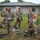 A military training or briefing session is taking place outdoors on a grassy field near a concrete block building. One soldier in the center appears to be leading a briefing or instruction session, gesturing with one hand while holding papers, while other soldiers in OCP (Operational Camouflage Pattern) uniforms listen from standing and seated positions. A civilian observer in civilian attire is visible in the background.