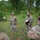 A group of U.S. Army soldiers in OCP (Operational Camouflage Pattern) uniforms are gathered outdoors in a grassy, wooded area. Two soldiers in the center appear to be conducting a briefing or after-action review, with one holding a green notepad and gesturing while the other holds a white document. A third soldier in the foreground is holding what appears to be a radio or communication device.