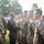 A military ceremony is taking place outdoors, with a senior service member walking down a line of uniformed personnel and shaking hands with individual soldiers. The personnel are wearing MARPAT (Marine pattern) digital camouflage uniforms, consistent with the United States Marine Corps. The scene appears to be a formal recognition or award ceremony conducted on a grass field with trees visible in the background.