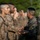 Two military personnel in U.S. Marine Corps MARPAT camouflage uniforms are engaged in a handshake, with one individual presenting what appears to be a wooden award or recognition item. They are surrounded by a formation of other uniformed Marines standing at attention in the background. The scene appears to be an outdoor military ceremony or award presentation.