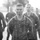 A black and white photograph showing a group of military personnel in camouflage uniforms, with the foreground subject visibly covered in mud or dirt on his face. The central figure wears a backpack/rucksack and appears fatigued or focused, suggesting completion of a physically demanding exercise. Several other uniformed individuals are visible in the background, slightly out of focus.