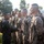 A military ceremony or inspection is taking place outdoors, with one service member shaking hands with another who is standing in a formed line. All personnel are wearing digital camouflage uniforms consistent with U.S. Marine Corps MARPAT pattern and are equipped with backpacks/rucksacks. The scene suggests a recognition event, graduation, or post-exercise acknowledgment.