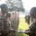 A military ceremony is taking place outdoors, likely an awards or recognition event. A female service member in MARPAT camouflage uniform is presenting items from a tray to another female service member standing at attention. Additional uniformed personnel are visible in formation in the background.