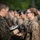 Two military personnel in U.S. Marine Corps MARPAT camouflage uniforms are engaged in what appears to be a formal ceremony, with one individual presenting or pinning something to the other. Both are wearing tactical gear including backpacks. A formation of additional military personnel is visible in the blurred background.