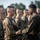 A military formation scene showing what appears to be a U.S. Marine Corps inspection or award ceremony. A senior Marine in MARPAT woodland camouflage is interacting closely with a junior Marine in desert MARPAT camouflage, possibly pinning an award or conducting a uniform inspection. Numerous other Marines stand in formation in the background.