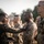 A military ceremony is taking place outdoors, where a senior service member in woodland MARPAT camouflage is pinning a decoration or device onto the collar/chest of a junior service member wearing desert MARPAT camouflage. The recipient stands at attention while other uniformed personnel are visible in the background in formation. The setting appears to be a formal military awards or recognition ceremony.