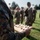 A U.S. Marine in MARPAT camouflage uniform holds a wooden presentation board engraved with the Eagle, Globe, and Anchor (EGA) emblem, with multiple metal pins or insignia arranged on it. The scene appears to be an outdoor military ceremony or formation on a grass field. Multiple Marines in uniform are visible standing in formation in the background.