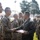A military awards ceremony is taking place outdoors, with one service member presenting a display board containing multiple medals or insignia to another. Both individuals are wearing Marine Corps MARPAT camouflage uniforms, and a formation of additional service members is visible in the background. The scene appears to be a formal recognition event conducted on a military installation.