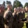 A military formation ceremony is taking place outdoors, with a line of uniformed service members standing at attention. An officer or senior NCO on the right appears to be presenting or handing something to a service member in the formation. The personnel are wearing MARPAT (Marine pattern) camouflage uniforms, consistent with U.S. Marine Corps dress.