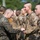 A military ceremony is taking place outdoors, where a senior service member appears to be presenting or pinning something to a junior service member. Both individuals are wearing U.S. Marine Corps MARPAT (Marine Pattern) camouflage uniforms. Multiple other Marines are standing in formation in the background.