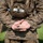A military personnel member in muddy camouflage uniform stands at parade rest, holding a small dark object (appears to be an insect or small creature) in their cupped hands behind their back. A name tape reading 'POOLAZ' is visible on a chest pouch. Other uniformed personnel are visible in the blurred background on a grassy field.