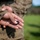 A person wearing U.S. Marine Corps MARPAT desert camouflage uniform is holding a small dark-colored Eagle, Globe, and Anchor (EGA) insignia pin in the palm of their hand. The EGA is the official emblem of the United States Marine Corps. The image is taken outdoors with a green, blurred background.