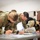 Combat aircraft integrate in Hawaii for exercise Sentry Aloha-21 Three military personnel in OCP (Operational Camouflage Pattern) uniforms are leaning over a table, writing on documents or forms. Two of the individuals are wearing black face masks, suggesting this was taken during the COVID-19 pandemic period. The setting appears to be an indoor administrative or processing area.