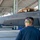 Combat aircraft integrate in Hawaii for exercise Sentry Aloha-21 A ground crew member wearing hearing protection and safety glasses observes an F-22 Raptor stealth fighter jet inside a covered aircraft shelter or hangar. The cockpit canopy is open and appears to have a golden/amber tint, with personnel visible in or around the cockpit area. At least two F-22 Raptors are visible under the shelter structure.