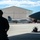 Combat aircraft integrate in Hawaii for exercise Sentry Aloha-21 A ground crew member wearing hearing protection stands in silhouette observing an F-15 Eagle fighter jet taxiing on the flight line at a military air base. A large aircraft hangar with a green roof is visible in the background, and the wing of another aircraft is partially visible on the right side of the frame. Ground support equipment, including a small wheeled cart, is positioned on the tarmac.