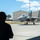 Combat aircraft integrate in Hawaii for exercise Sentry Aloha-21 A military ground crew member wearing hearing protection observes an F-15 Eagle fighter jet taxiing on the flight line at what appears to be a U.S. Air Force base. The aircraft is in a gray paint scheme and appears to be carrying external fuel tanks or ordnance. Military hangars with green roofing are visible in the background under a partly cloudy sky.