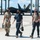 Combat aircraft integrate in Hawaii for exercise Sentry Aloha-21 Three military personnel wearing black face masks walk across an airfield tarmac in what appears to be a tropical location. Behind them, a stealth fighter jet — consistent with an F-22 Raptor — is visible inside or near a large hangar structure. The setting suggests a U.S. Air Force base, likely in Hawaii based on the palm trees and terrain.