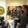Combat aircraft integrate in Hawaii for exercise Sentry Aloha-21 A group of military personnel wearing olive green flight suits and face masks are gathered in what appears to be a squadron operations room or briefing area. They are standing at or near a counter with computers, appearing to attend a briefing or receive information. A squadron patch/emblem is visible on the wall behind them, along with what appears to be a unit insignia reading '19th Fighter SQ' or similar designation.