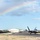 Combat aircraft integrate in Hawaii for exercise Sentry Aloha-21 Two F-22 Raptor stealth fighter jets are taxiing on a runway at what appears to be a military airbase, likely in Hawaii based on the tropical vegetation and distinctive architecture. A vivid rainbow arcs across a partly cloudy blue sky in the background, creating a striking visual contrast with the military aircraft. Military hangars and infrastructure are visible along the flight line.