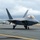 Combat aircraft integrate in Hawaii for exercise Sentry Aloha-21 An F-22 Raptor stealth fighter jet is taxiing on a runway, with at least one additional aircraft visible in the background. The aircraft displays tail code 'HH' indicating assignment to Hawaii (Hickam Air Force Base / Joint Base Pearl Harbor-Hickam). Mountains, urban development, and overcast skies are visible in the background.