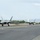 Combat aircraft integrate in Hawaii for exercise Sentry Aloha-21 Three F-22 Raptor stealth fighter jets are taxiing in formation on a military airfield runway. The aircraft appear to be preparing for takeoff or returning from a mission, moving in a single-file line along the taxiway. The setting appears to be a coastal military installation, likely in Hawaii based on the terrain and vegetation.