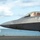 Combat aircraft integrate in Hawaii for exercise Sentry Aloha-21 A close-up photograph of a stealth fighter jet, identifiable as an F-22 Raptor, taxiing on a runway near a coastal waterway. A pilot in full flight gear is visible through the canopy, appearing to wave or gesture. The aircraft displays a name placard on the fuselage and is photographed against a partly cloudy sky.