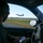 Combat aircraft integrate in Hawaii for exercise Sentry Aloha-21 A military personnel member wearing a flight suit and face mask sits inside a Chevrolet vehicle near an airfield runway, holding a smartphone. Through the open window, a military jet aircraft — appearing to be an F-15 — is visible taking off or landing on the runway. The scene suggests airfield operations monitoring or coordination activity.