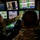 Two military personnel in camouflage uniforms are seated at a multi-screen ground control station, likely operating or monitoring an unmanned aerial vehicle (UAV/drone). One operator is pointing at a central monitor displaying what appears to be a camera feed from an aircraft, while multiple screens show maps, telemetry data, and surveillance imagery. The environment is a darkened control room with numerous displays showing real-time operational data.