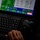 The image shows a ground control station interface with a specialized keyboard, trackball, and joystick controls visible in the foreground. A monitor displays what appears to be a UAV (unmanned aerial vehicle) or drone control software interface with multiple data readouts, status indicators, and mission control buttons. A human hand is visible operating the keyboard in low-light conditions.