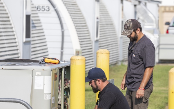 Two male technicians are servicing an HVAC/air conditioning unit outdoors near a row of metal storage or utility buildings. One technician is crouching down inspecting the open panel of the unit while the other stands and observes. The setting appears to be a commercial or industrial facility with yellow safety bollards lining a concrete walkway.