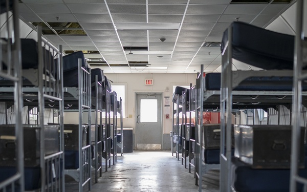 The image shows a large, currently unoccupied dormitory-style room filled with rows of metal bunk beds equipped with blue mattresses. The facility appears institutional in nature, consistent with a detention center, shelter, or military barracks. The space shows signs of wear, including stained concrete flooring and a damaged ceiling tile.
