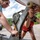 Pittsburgh District hosts motorboat training Two young women are working together to secure or release a boat on a trailer, using a manual winch mechanism and chain hook. The scene takes place on a boat ramp or gravel launch area on a sunny day. Both individuals are wearing life vests, suggesting they are preparing to launch or retrieve the boat.