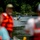 Pittsburgh District hosts motorboat training A waterway scene showing people observing boats on a calm river or lake surrounded by dense green trees. In the foreground, out-of-focus observers are visible, including a person wearing an orange life vest. Two motorboats are visible in the mid-ground with people aboard.
