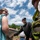 Pittsburgh District hosts motorboat training A group of people are gathered outdoors near what appears to be a boat, likely at a boat launch or waterway access point. A man in a black long-sleeve shirt and cap appears to be instructing or conversing with others, including a young girl in a teal top and a person wearing a yellow life vest. The setting is a sunny day with blue skies and green tree-covered hills in the background.