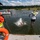 Pittsburgh District hosts motorboat training A water safety training or rescue drill is taking place on a lake, with multiple participants wearing life vests jumping or already in the water. A person in a yellow high-visibility vest and orange life preserver observes from a dock or platform, while another person in a reflective vest is mid-jump into the water. A Park Ranger boat with personnel is visible in the background overseeing the activity.