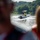 Pittsburgh District hosts motorboat training A motorboat with two occupants is navigating a river, creating a visible wake behind it. The scene is photographed from another vessel, with two blurred figures in the foreground framing the shot. The background features lush green tree-covered hills under bright daylight conditions.