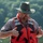 Pittsburgh District hosts motorboat training A middle-aged man wearing a red life vest and olive-colored wide-brim hat stands near a body of water with green, tree-covered hills in the background. He appears to be demonstrating or adjusting his personal flotation device (PFD), with both hands gesturing toward the vest. The image is well-lit with natural outdoor lighting suggesting a warm, sunny day.