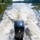 Pittsburgh District hosts motorboat training The image shows a rear-facing view from a motorboat traveling along a calm river or waterway, with a prominent black outboard motor (Mercury brand) visible in the foreground. A wide, foamy white wake trails behind the boat, spreading across the greenish water. Tree-lined banks with dense green foliage frame both sides of the waterway.