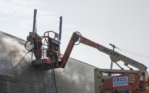 A JLG 340AJ articulating boom lift, rented from United Rentals, is being used to perform maintenance or cleaning work on the exterior wall of a Quonset hut-style corrugated metal building. A worker in the lift basket appears to be pressure washing or applying a treatment to the building's surface, as evidenced by visible mist or spray. Safety cones, including an orange 'Sidewalk Closed' cone and green cones, are placed on the ground nearby.